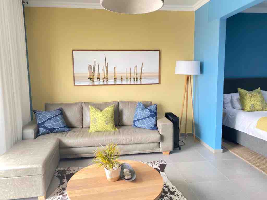 A modern living area features a light-colored sofa with patterned cushions, accompanied by a round wooden coffee table. A decorative plant is positioned on the table. A painting of weathered wooden posts hangs on the wall, complementing the two-tone color scheme of yellow and blue.