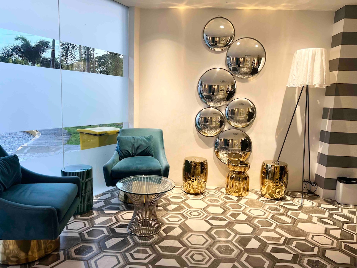 A stylish lounge area features modern furniture, including two teal armchairs and a round glass table. A set of mirrored wall balls adds visual interest, while golden side tables complement the geometric patterned floor. Natural light filters through a large window, enhancing the space.
