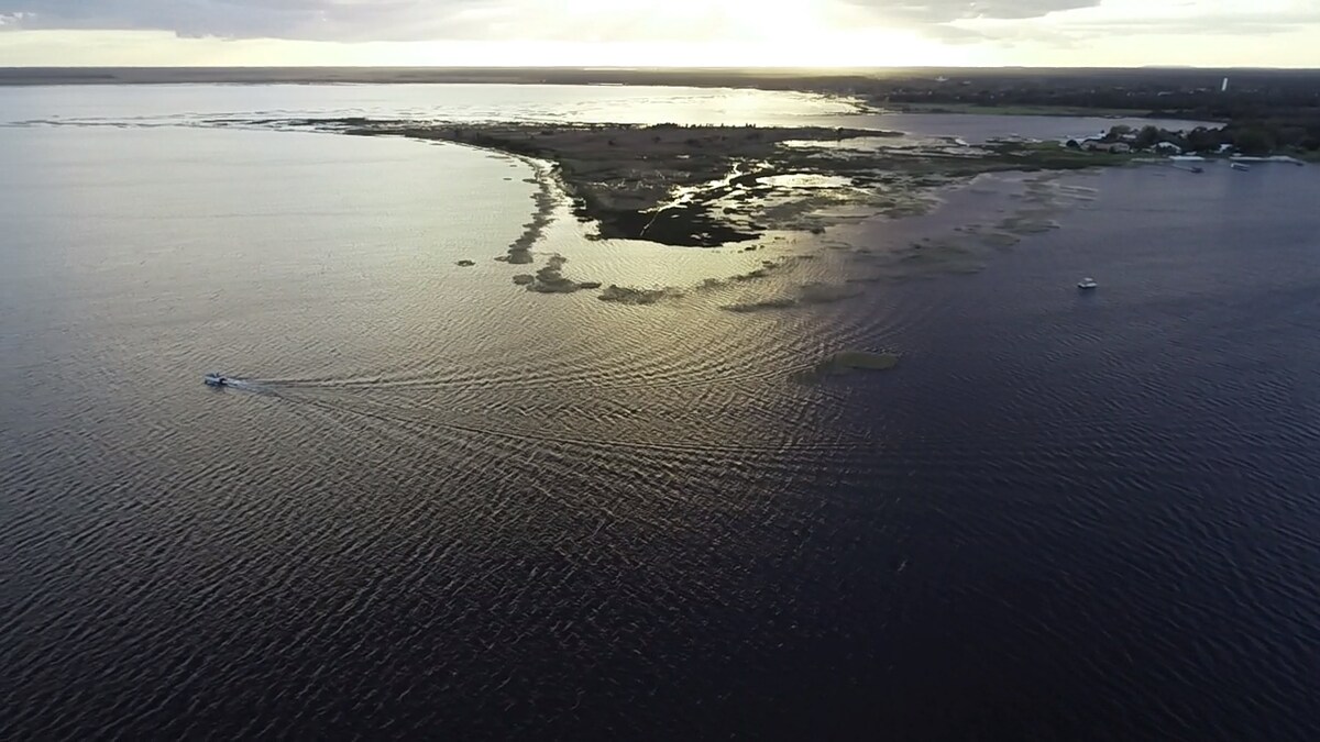 Aerial view of Crooked Lake displaying its expansive waters reflecting the soft glow of sunset. The shoreline features lush vegetation and small islands, with boats gently traversing the smooth surface, creating ripples. The scene offers a blend of tranquility and natural beauty.