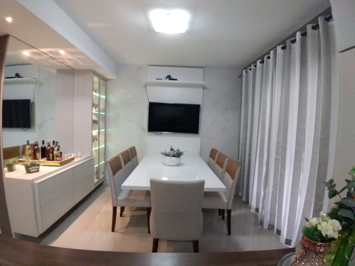 A modern dining area features a large white table surrounded by plush chairs, suitable for gatherings. A wall-mounted television is positioned above, and a decorative bar with assorted bottles adds a touch of elegance. Soft, natural light filters through sheer curtains.