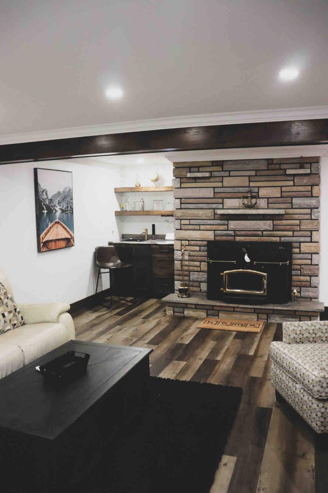 A cozy living area features a stone fireplace with a wood-burning stove and a wooden mantel. Comfortable seating is arranged around a black coffee table, and a shelf displays decorative items. Soft lighting creates a relaxed ambiance throughout the space.