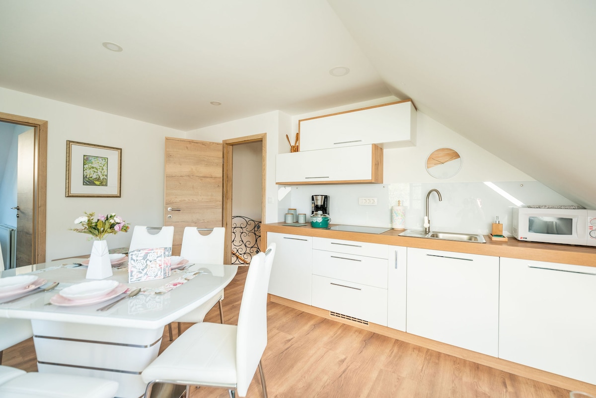 A modern kitchenette is displayed, featuring white cabinetry with wood accents. A dining table is set with tableware, complemented by a variety of appliances including a coffee maker, microwave, and sink. Natural light brightens the space through a slanted ceiling.