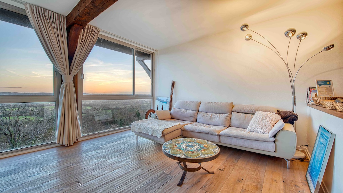 A spacious living area features a comfortable sofa accompanied by a patterned coffee table. Large windows provide a panoramic view of the surrounding nature, with soft light filtering through sheer curtains. The warm wooden floor complements the serene atmosphere.