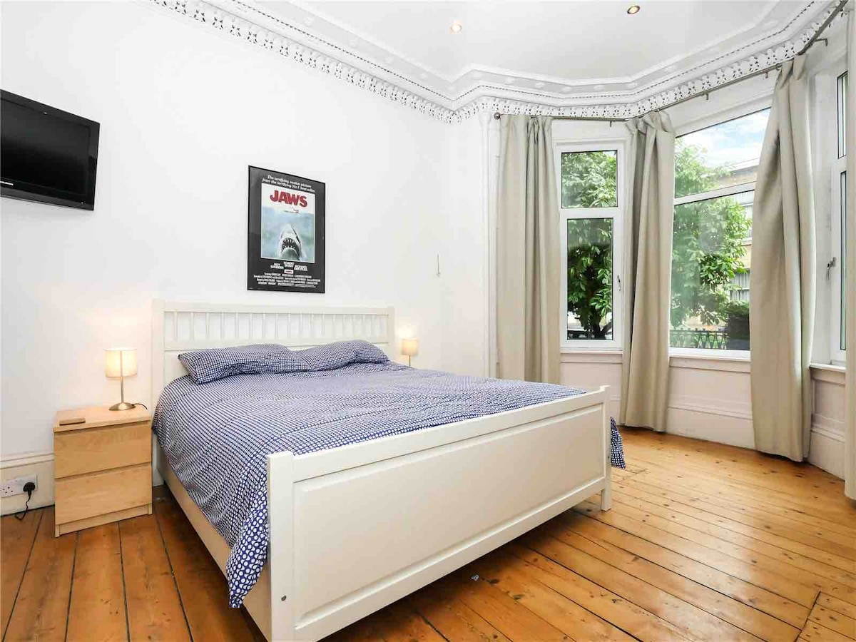 A spacious bedroom features a large bed with a blue-patterned coverlet and light-colored wooden flooring. Natural light streams through large bay windows, which are framed by neutral drapes. A small nightstand with an illuminated lamp and a wall-mounted television is also visible.