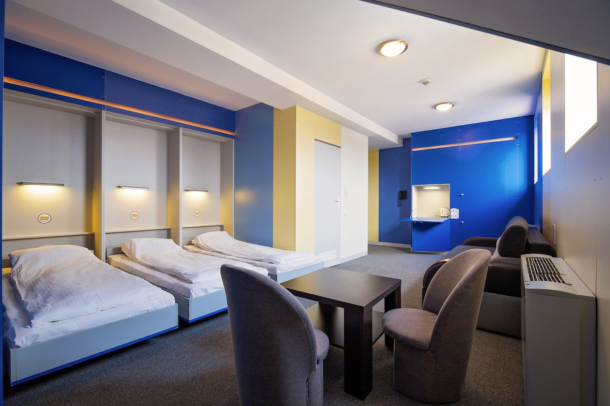 A spacious family-friendly room features two separate beds with simple white bedding. A small table is positioned centrally, surrounded by two chairs. A kitchenette area is present along one wall, while natural light enters through a high window, highlighting the blue and yellow color scheme.