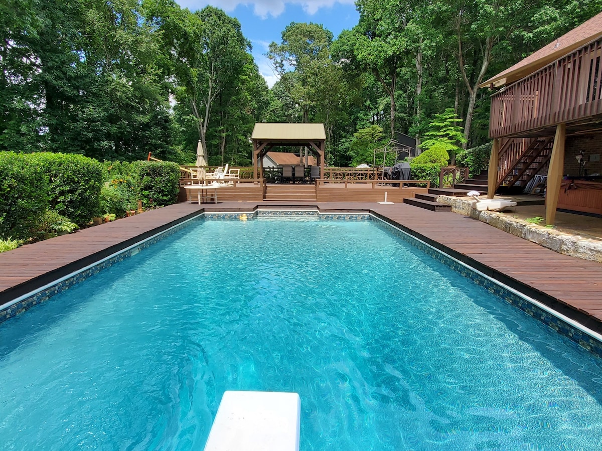 A clear, blue pool is surrounded by lush greenery and wooden decking. Lounge chairs are arranged in a shaded area with a gazebo in the background. A spacious deck is visible, providing an inviting space for relaxation and outdoor gatherings.