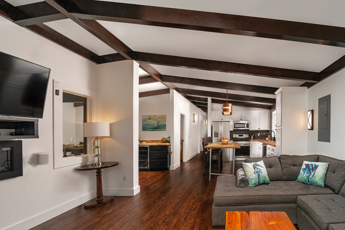 An open living space features exposed wood beams overhead and a modern sofa adorned with decorative pillows. A mounted flat-screen TV and electric fireplace are positioned on one wall, while the kitchen with stainless steel appliances is visible in the background.