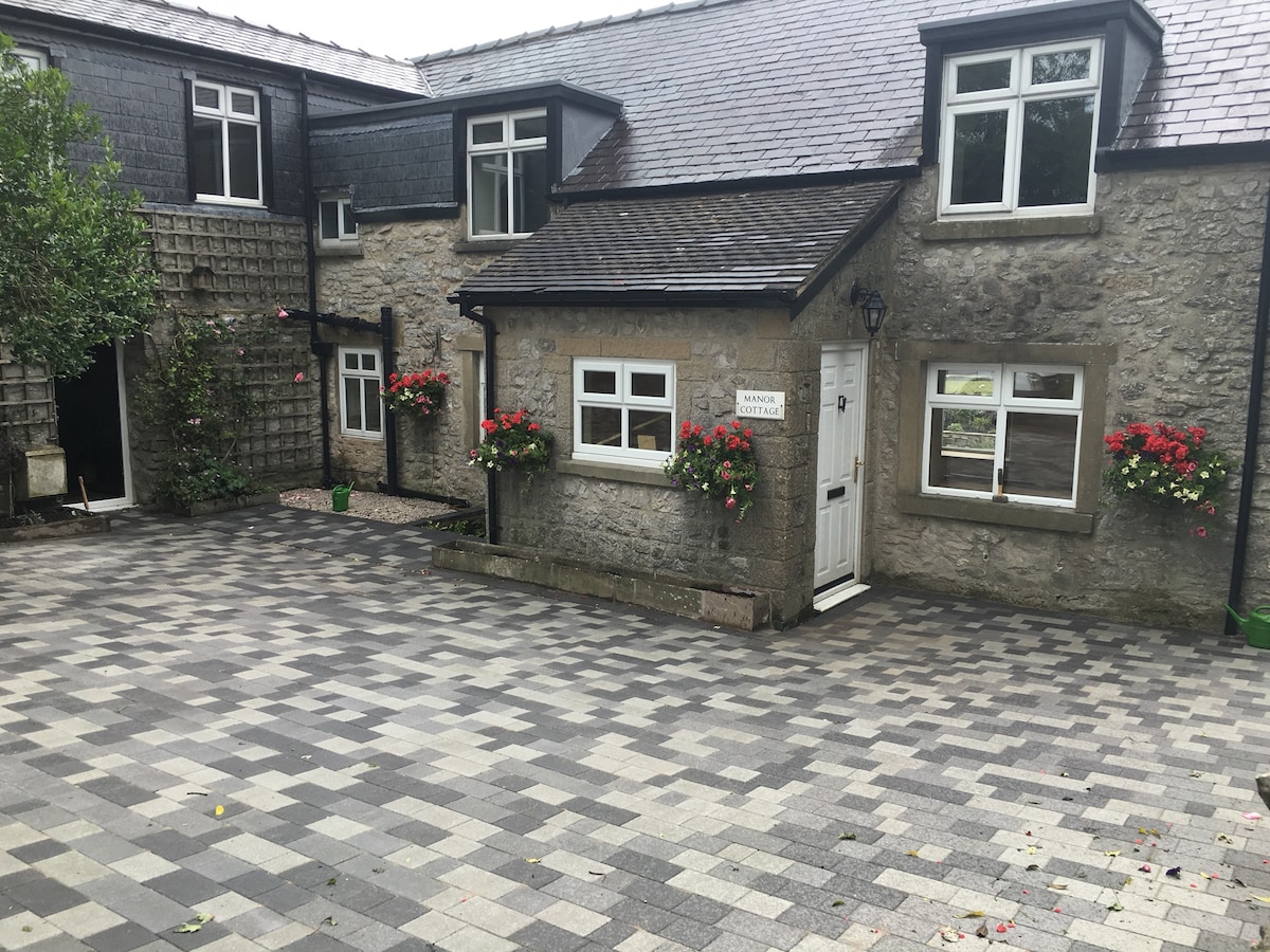 The exterior of Manor Cottage showcases a blend of traditional stone and modern architecture. Flower boxes in vibrant colors adorn the windows, while a well-maintained paved courtyard offers ample parking space. The surrounding greenery adds an inviting touch to the overall setting.