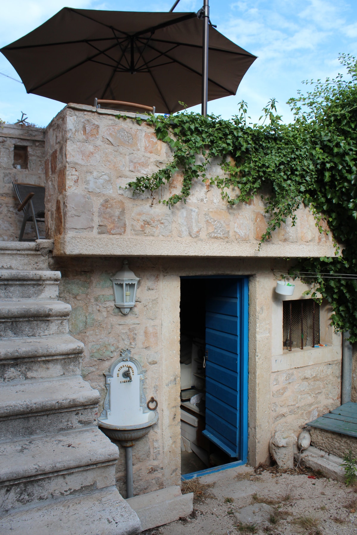 A sturdy stone structure showcases a vibrant blue door that opens into the interior. A patio umbrella provides shade above the entrance, while climbing greenery adds a touch of nature. Stairs lead to the upper level, enhancing the building's charm.