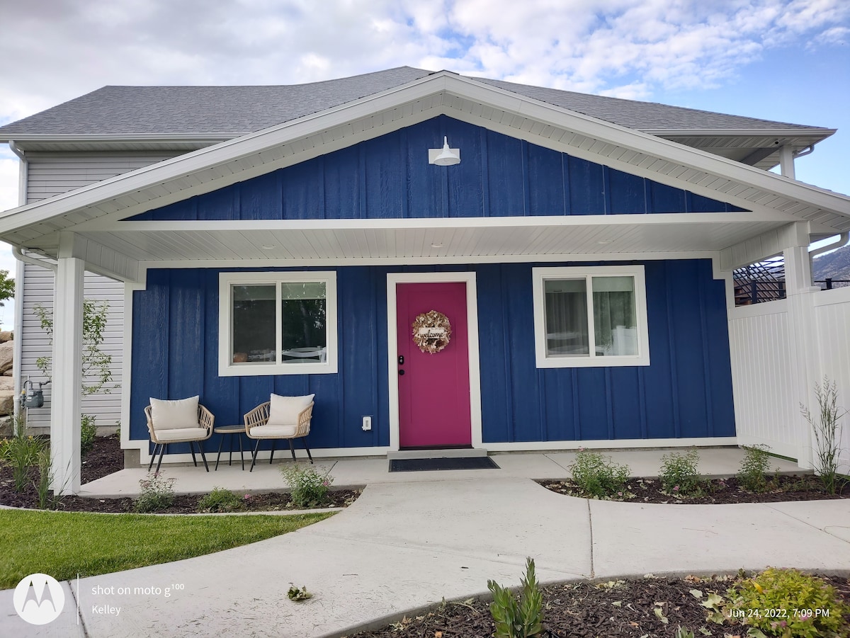 The exterior of a charming blue home is showcased, featuring a welcoming pink front door. Flanking the entrance are two light-colored chairs. A neatly manicured path leads to the door, with green landscaping enhancing the property's inviting appearance.