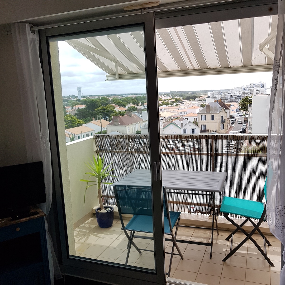 A balcony setting is presented with a small table and two folding chairs, framed by a protective railing. A view of the surrounding neighborhood and distant greenery is visible through the sliding glass door. Natural light enters, enhancing the space's bright atmosphere.