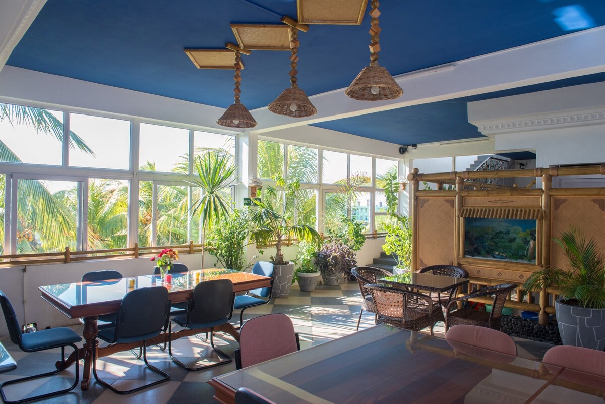 A bright communal area is featured, with large windows allowing natural light to illuminate the space. Tropical plants are positioned throughout, enhancing the atmosphere. Various seating options, including tables and chairs, are provided for guests, creating an inviting area for social interactions.