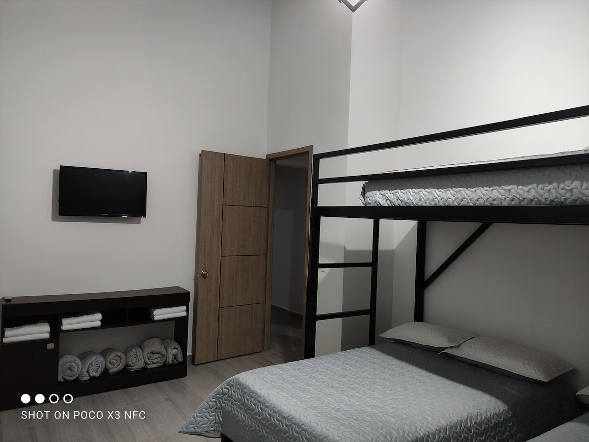 A simple sleeping area features a dual-level bunk bed and a spacious bed with soft bedding. A flat-screen television is mounted on the wall, and a wooden cabinet displays neatly folded towels. The room is brightly lit, providing a comfortable and inviting atmosphere.