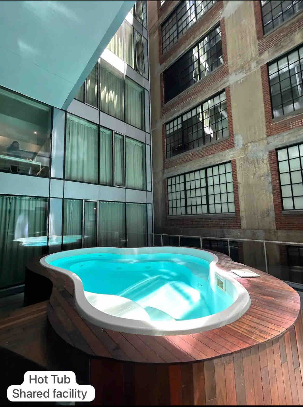 A shared hot tub is situated in a serene courtyard. The wooden deck encircles the tub, while large, industrial-style windows rise in the background, allowing natural light to fill the space. The calm water reflects the surrounding materials.