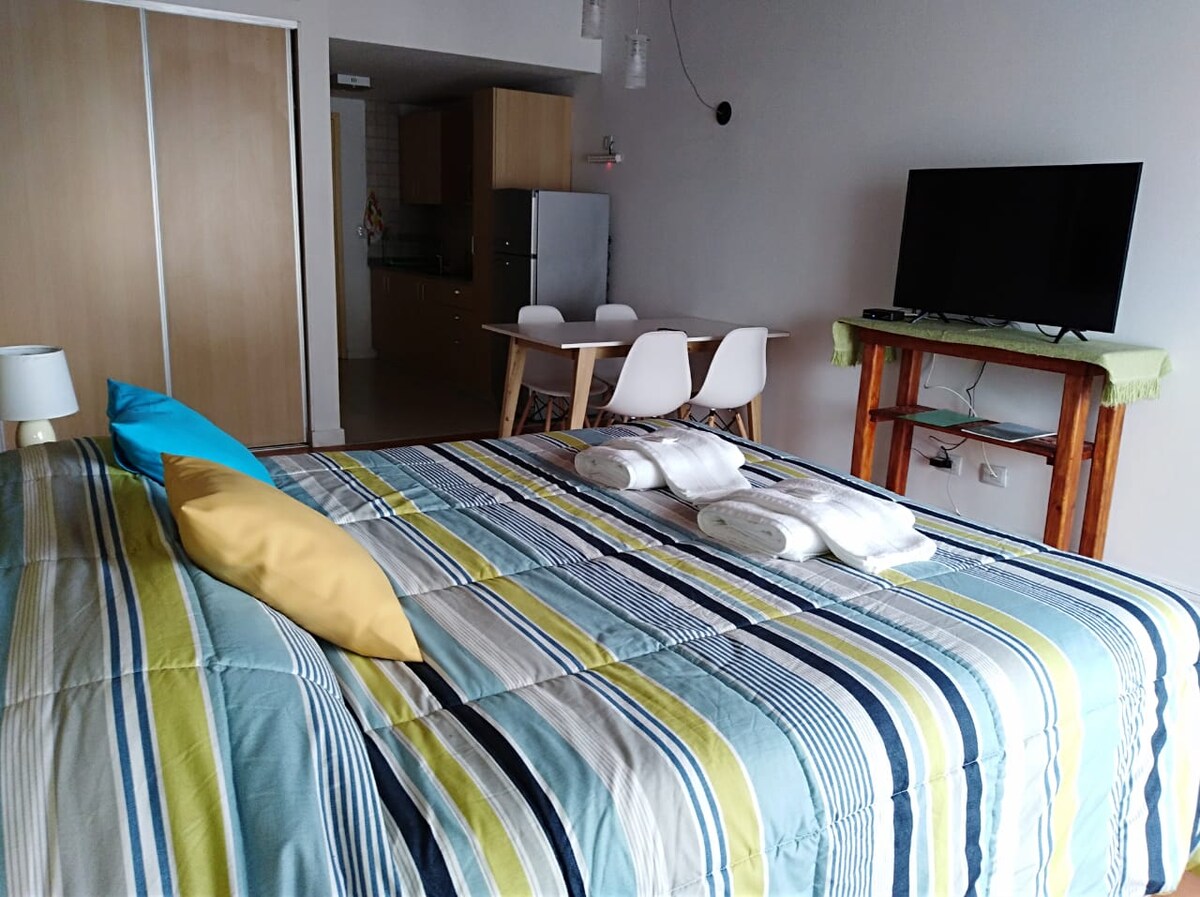 A spacious sleeping area features a striped comforter layered on a large bed, complemented by colorful throw pillows. A dining space, equipped with a wooden table and white chairs, is visible in the background, along with a flat-screen television mounted on the wall.