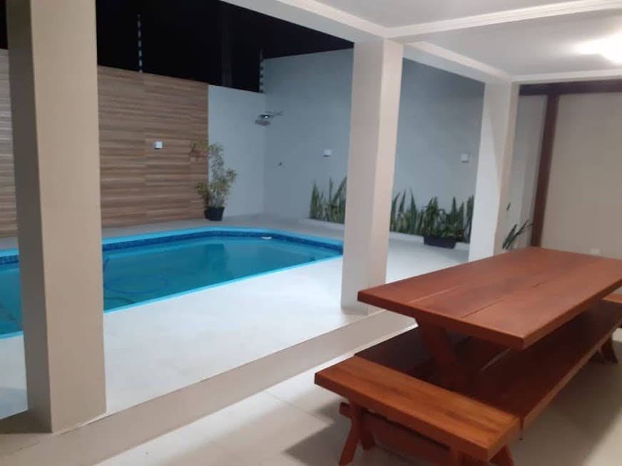 A well-lit outdoor area features a rectangular pool surrounded by tile flooring. A wooden dining table and benches are positioned near the pool, with plants lining the space partially in view, contributing to a relaxed environment.