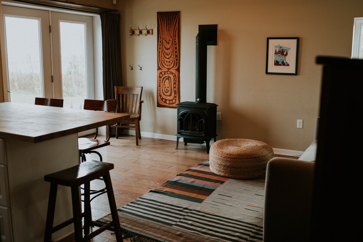 An open living area features a cozy seating arrangement with a plush pouf and a warm wood stove. Nearby, a dining area is highlighted by wooden chairs and a high table, while a vibrant area rug adds texture to the space.