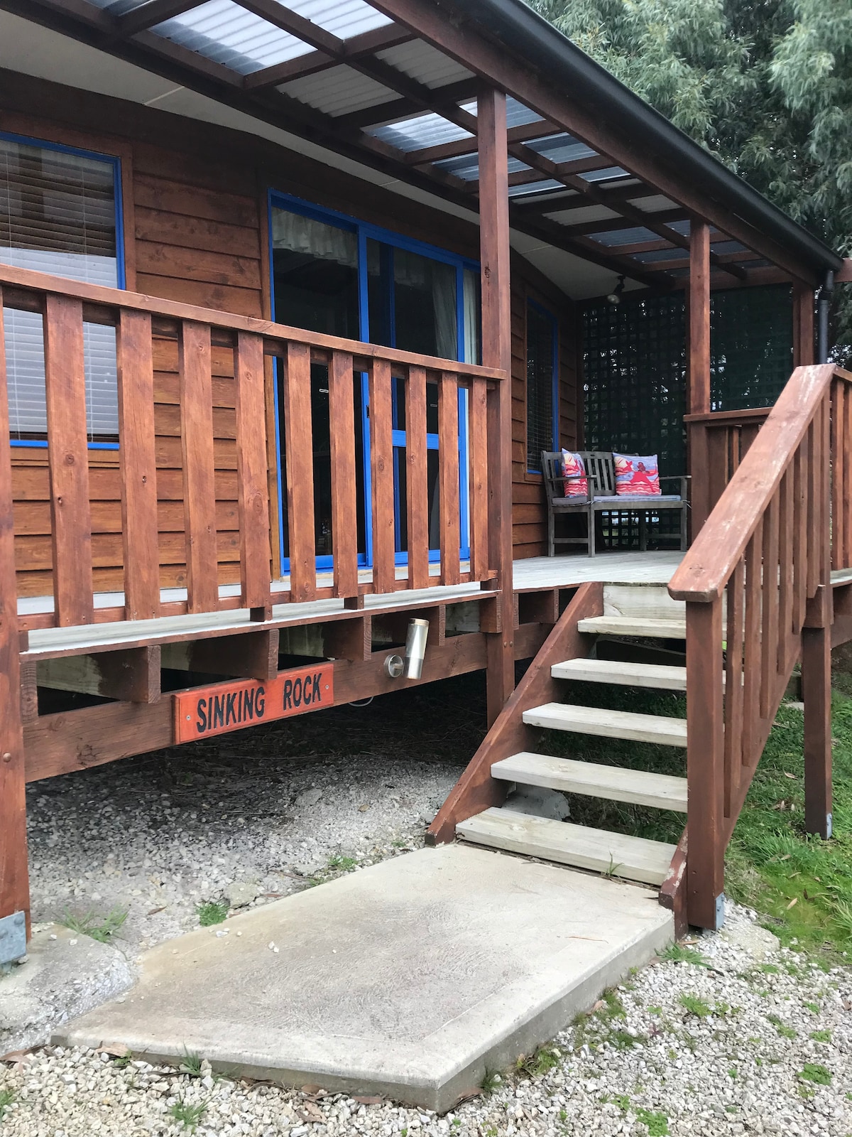 The entrance of the cozy cabin is highlighted by a wooden porch and steps leading up to the front door, featuring a sign that reads 'SINKING ROCK.' The porch includes seating for relaxation, surrounded by greenery.