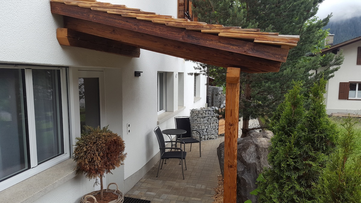 An outdoor seating area is situated beneath a wooden overhang, featuring a small round table and two chairs. Lush greenery and decorative plants are present, enhancing the peaceful environment. The entrance to the building is visible, complemented by a stone pathway.
