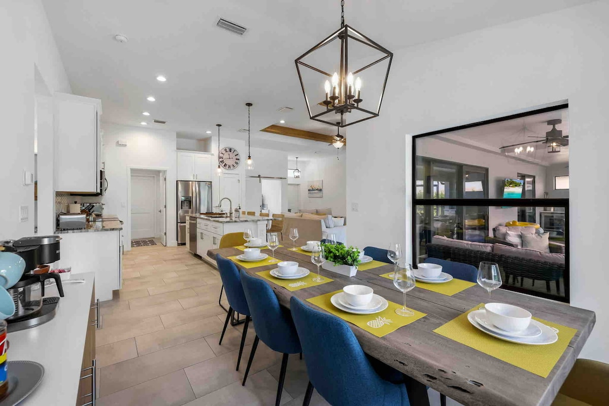 A modern dining area features a long wooden table set for eight, adorned with yellow placemats and white dishware. The space is illuminated by a stylish chandelier. The open layout connects to a bright kitchen, showcasing stainless steel appliances and a cozy living area visible through large windows.
