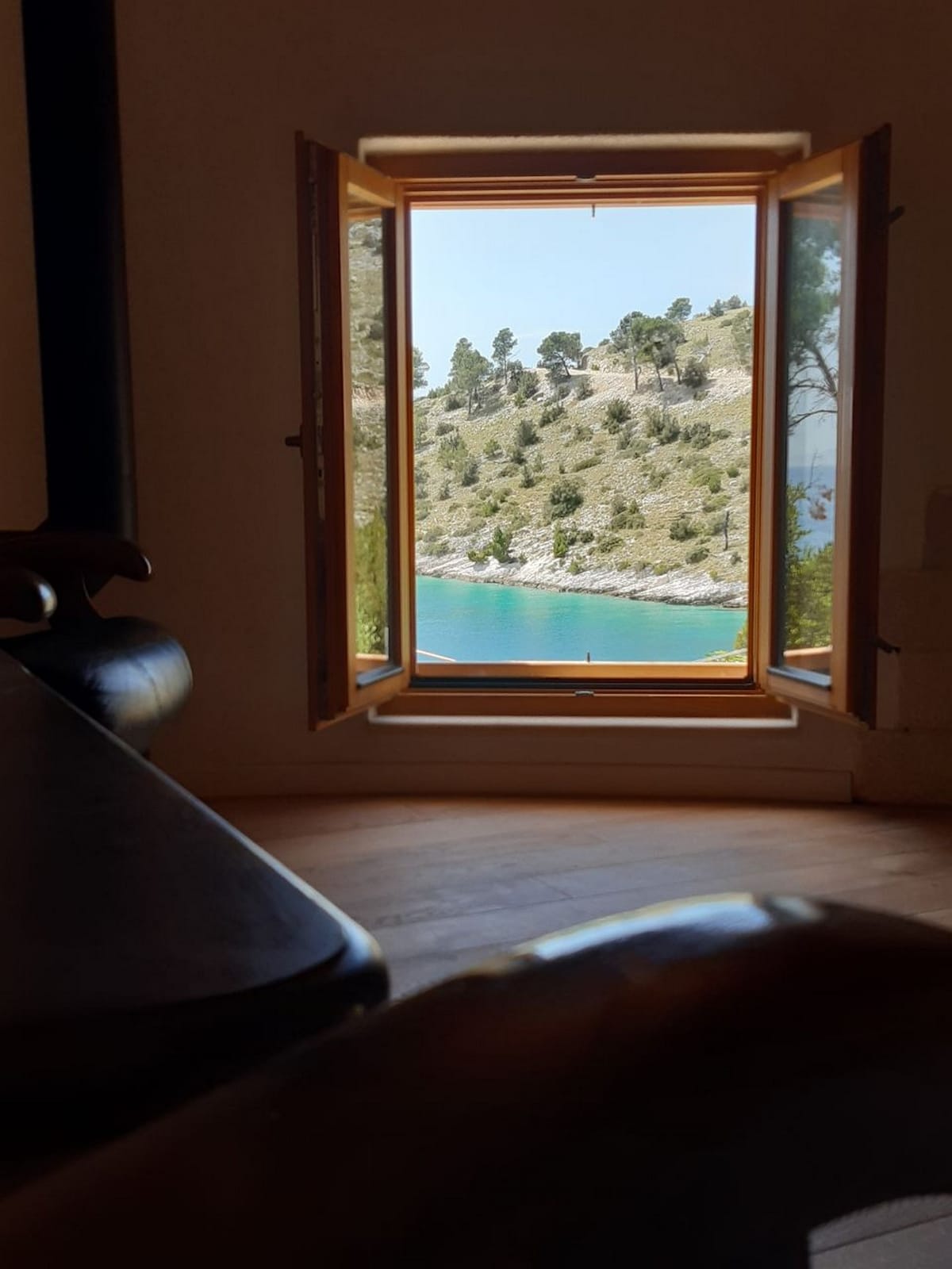 A view is presented from an open window, showcasing lush green hills sloping down to a serene blue bay. Natural light is seen flooding into the room, enhancing the tranquil atmosphere created by the landscape.