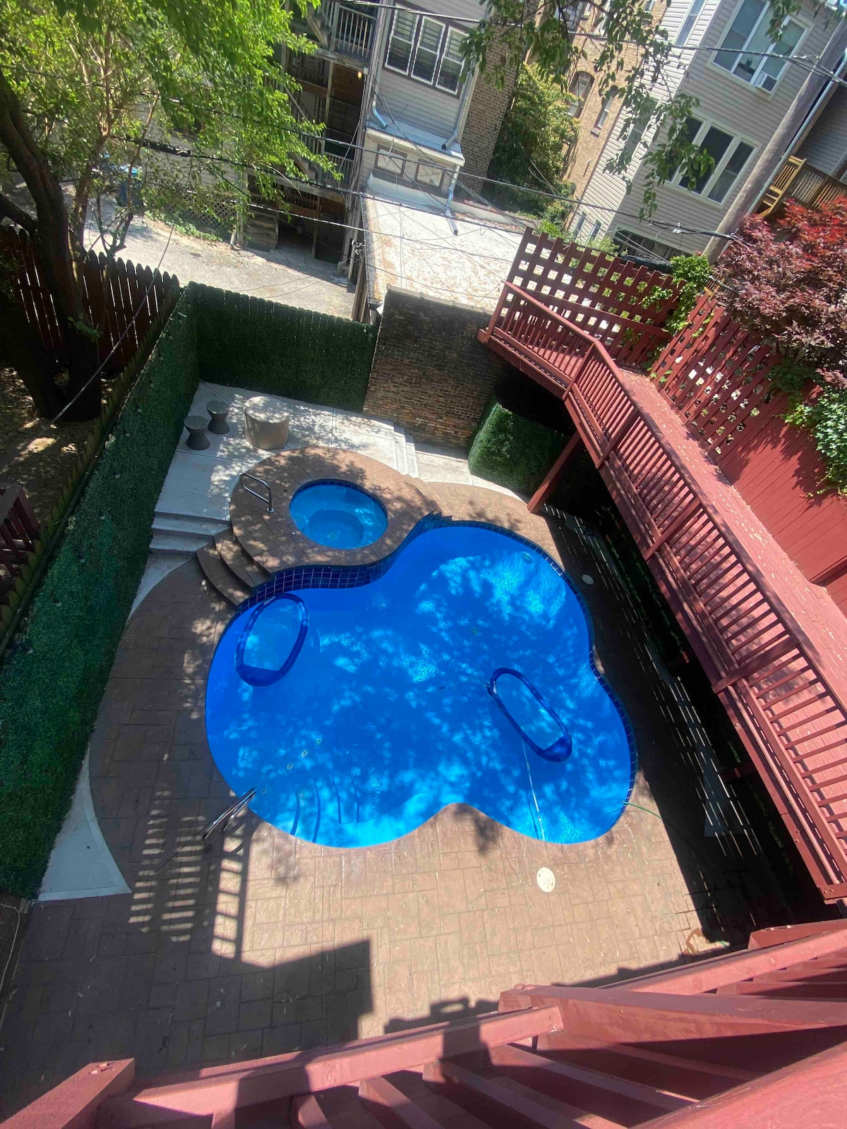 An aerial view of the outdoor area reveals a blue pool shaped comfortably for relaxation, accompanied by a smaller adjacent hot tub. Surrounding greenery and wooden decking provide a private setting, with ample space for lounging and enjoying the outdoors.
