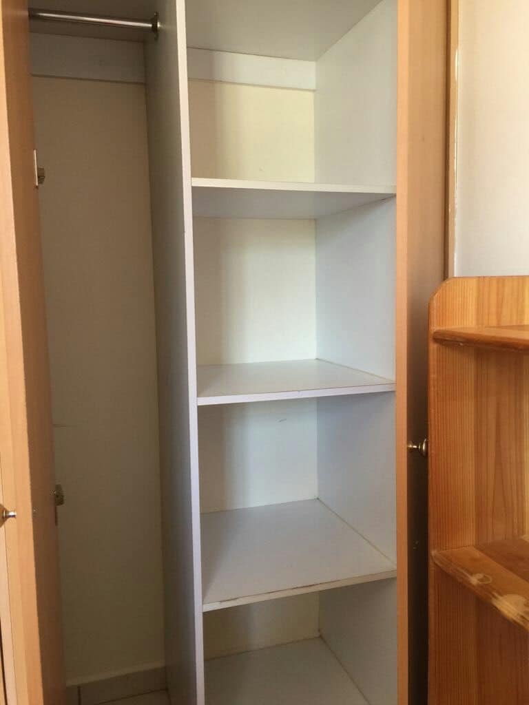 A built-in closet is shown with several empty shelves. The interior is minimally decorated, providing ample space for storage. The closet door is partially open, allowing a glimpse of the light-colored shelving against a plain backdrop.