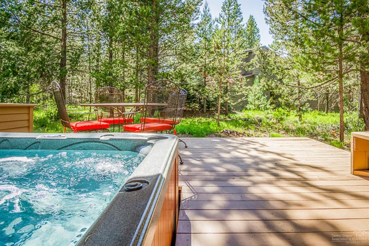 A hot tub is positioned on a wooden deck, surrounded by green pine trees. A dining table with four chairs is visible in the background, enhancing the outdoor space. Sunlight filters through the trees, casting playful shadows on the deck.