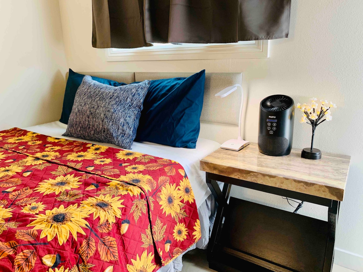 A neat sleeping area with a comfortable bed covered in a floral quilt is shown. Two pillows provide support. A small bedside table with a lamp is visible, along with a portable heater and a decorative plant, enhancing the cozy environment.