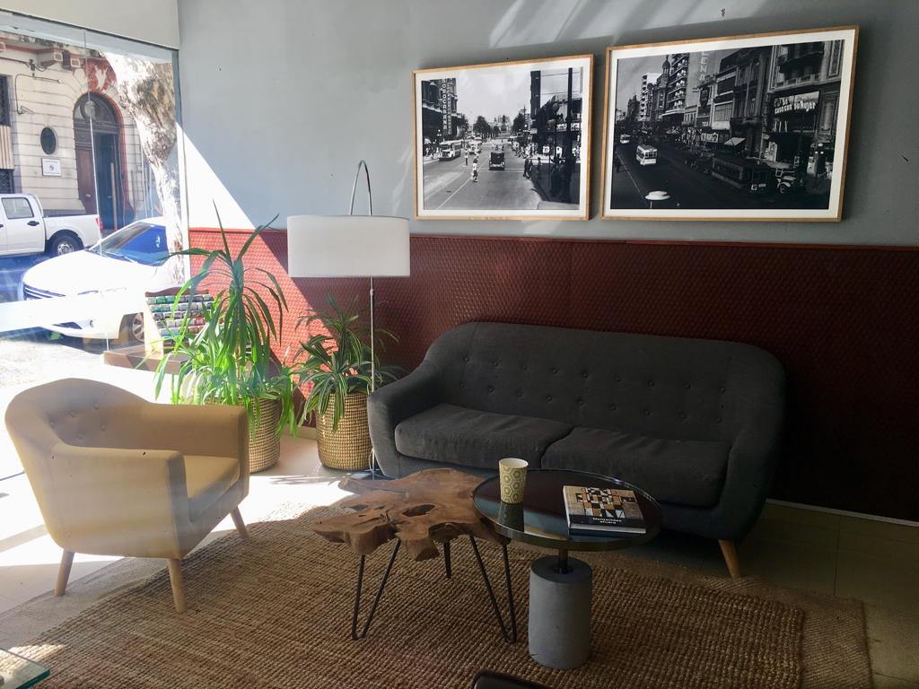 A welcoming lounge area features a gray sofa and a light-colored armchair, positioned on a woven rug. A round coffee table stands in front, topped with a magazine and a cup. Large framed black-and-white photographs decorate the wall, complemented by potted plants.