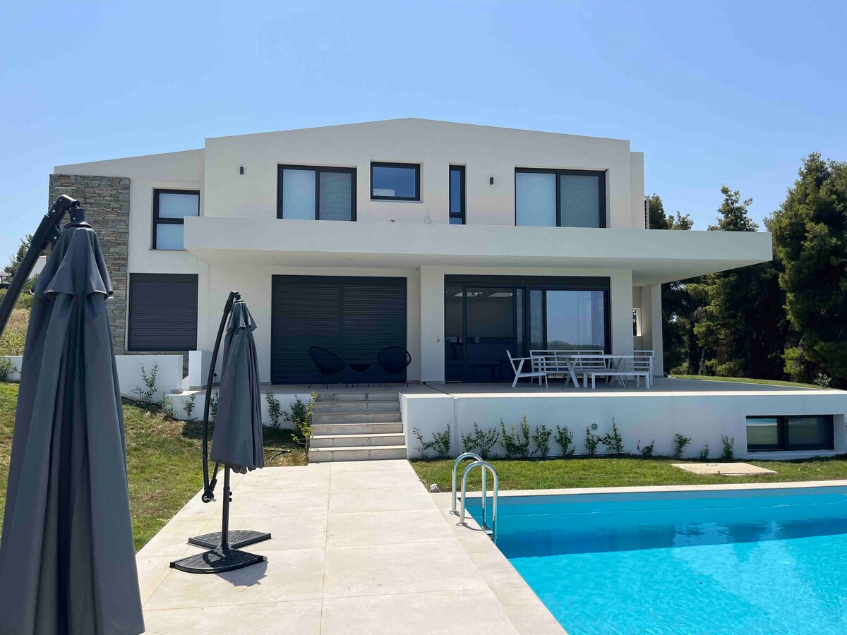 A modern villa features a sleek design and large windows, allowing natural light to fill the spaces. The exterior includes a spacious terrace with outdoor seating and a private pool, surrounded by well-maintained gardens that blend into the natural landscape.
