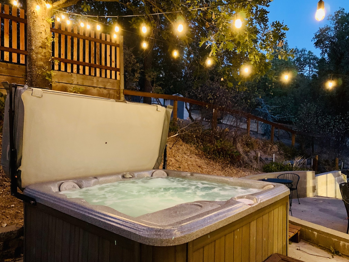 A hot tub is seen nestled among trees, with evening lights gently illuminating the area. The hot tub cover is partially raised, revealing steaming water inside. A natural landscape provides a serene backdrop, enhancing the relaxation experience.