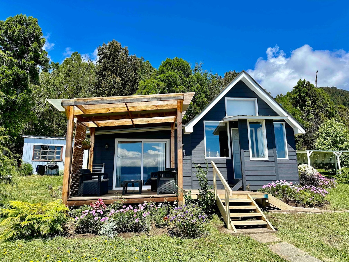 A spacious cabin with a welcoming covered terrace is depicted, featuring large glass doors that reveal scenic views. Colorful flowers line the pathway, leading to the entrance, while lush greenery surrounds the property, enhancing the natural setting.