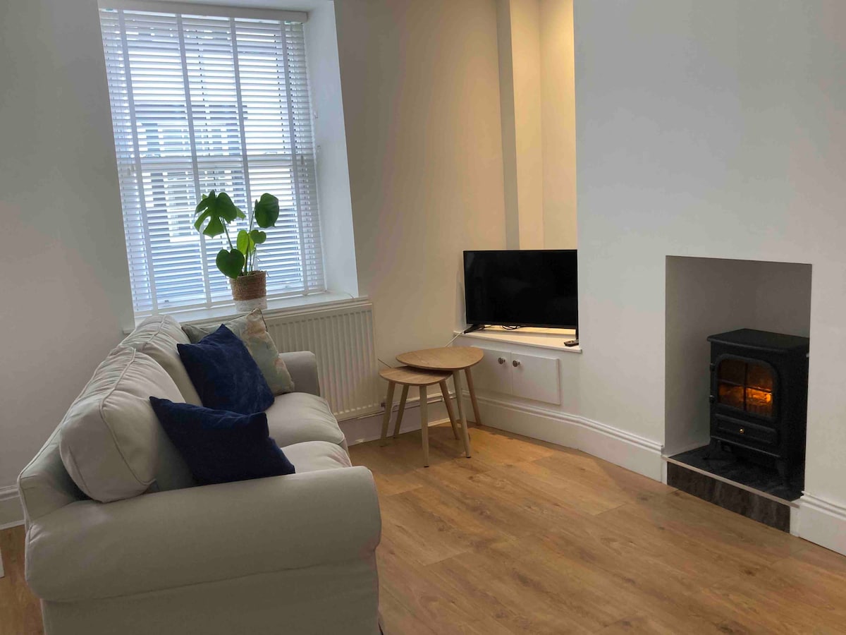 A cozy living area features a light-colored sofa with dark cushions positioned near a window with blinds. A television is placed on a stand across from a small, round table, and a simple electric fireplace is set into the wall, providing warmth.