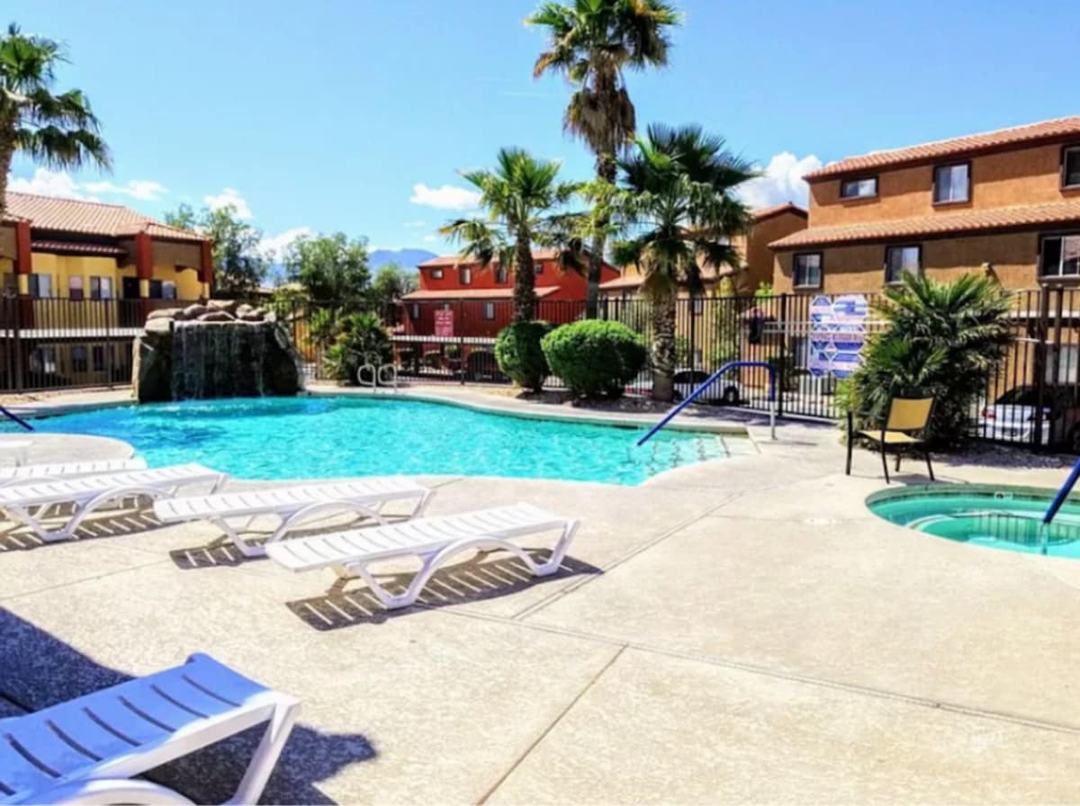 The outdoor pool area features clear water, surrounded by lounge chairs for sunbathing. Nearby, a hot tub is positioned for relaxation. Palm trees provide shade, with a landscaped area including bushes and a rock formation. The fenced area enhances privacy and security.