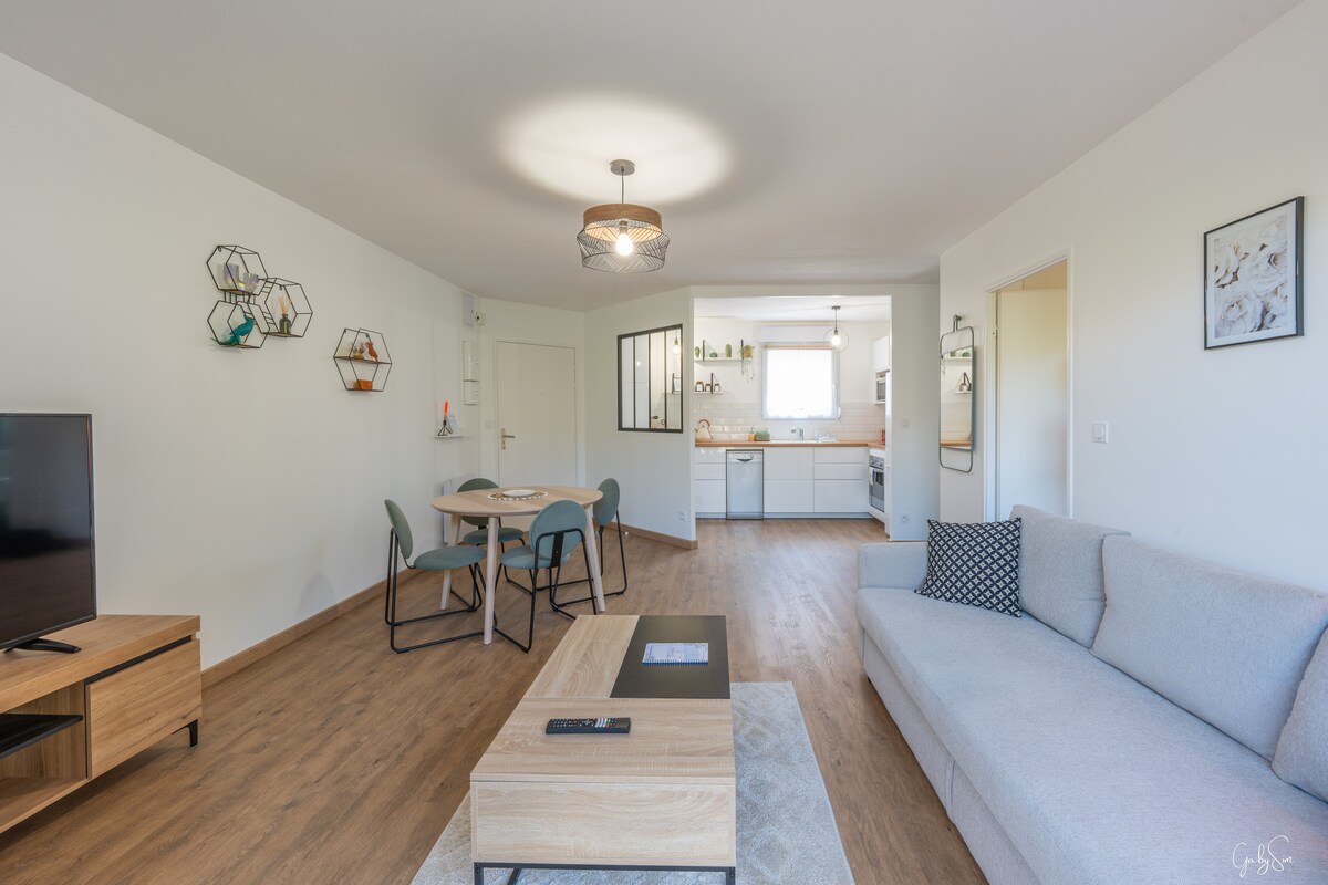 A welcoming living area is highlighted by a light-colored sofa and a modern coffee table. A round dining table with four chairs is positioned nearby. The kitchen, visible in the background, features clean lines and ample natural light from a window.