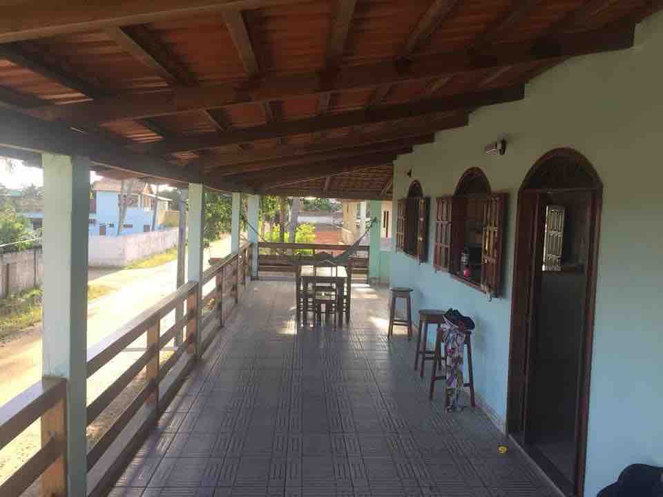 A spacious outdoor veranda is covered and features wooden railings. A set of four chairs and a table are arranged for dining. Natural light filters through multiple arched windows, highlighting the tiled floor. Lush greenery can be seen along the pathway outside.