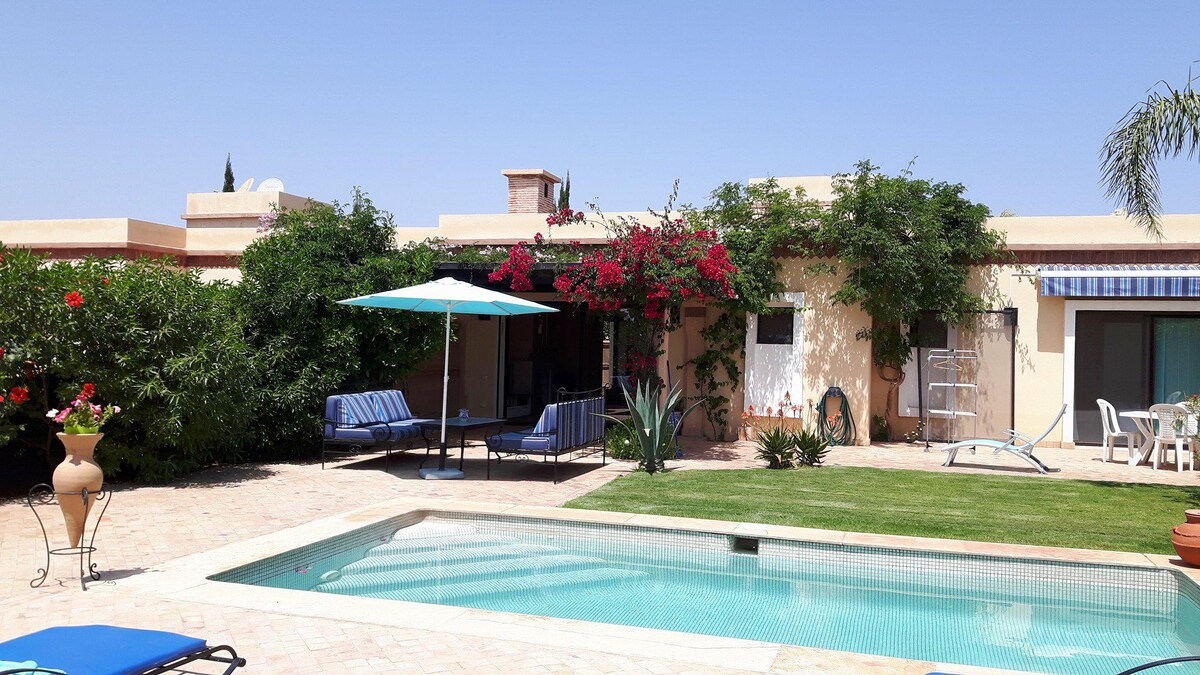 A modern villa is set within a landscaped garden, featuring a private pool and shaded seating area. Bougainvillea climbs the exterior walls, adding vibrant color. Comfortable lounge chairs are positioned by the pool, providing a relaxing outdoor space. Clear blue skies complete the serene ambiance.