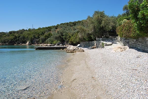 A serene beach scene is presented, showcasing a shoreline of smooth pebbles transitioning to fine sand. Clear blue waters gently lap at the shore, while lush greenery borders the beach. A small wooden pier extends into the water, offering a peaceful view of the surrounding landscape.