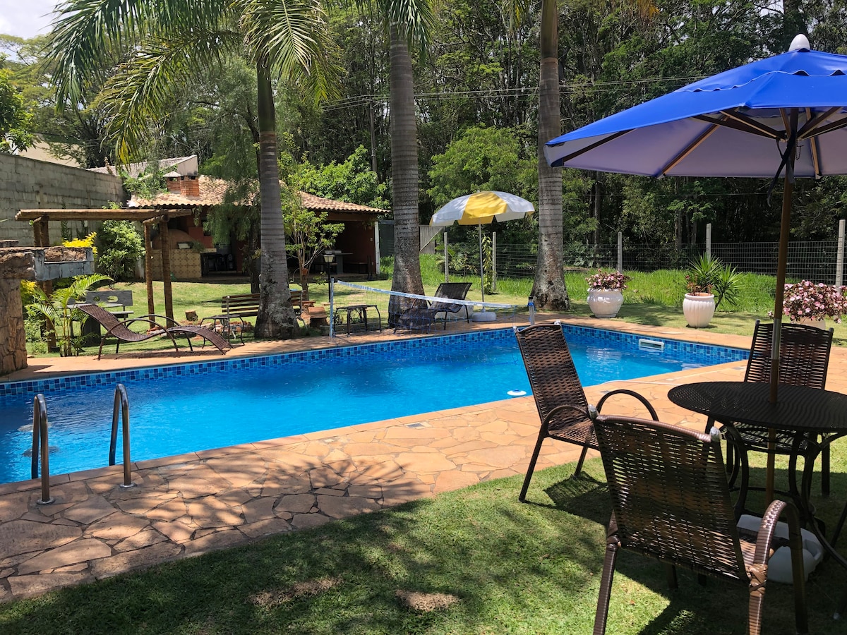An outdoor area features a rectangular pool, surrounded by palm trees and a manicured lawn. Lounge chairs are positioned nearby, along with a table shaded by an umbrella. A rustic gazebo and additional seating are visible in the background, enhancing the natural setting.
