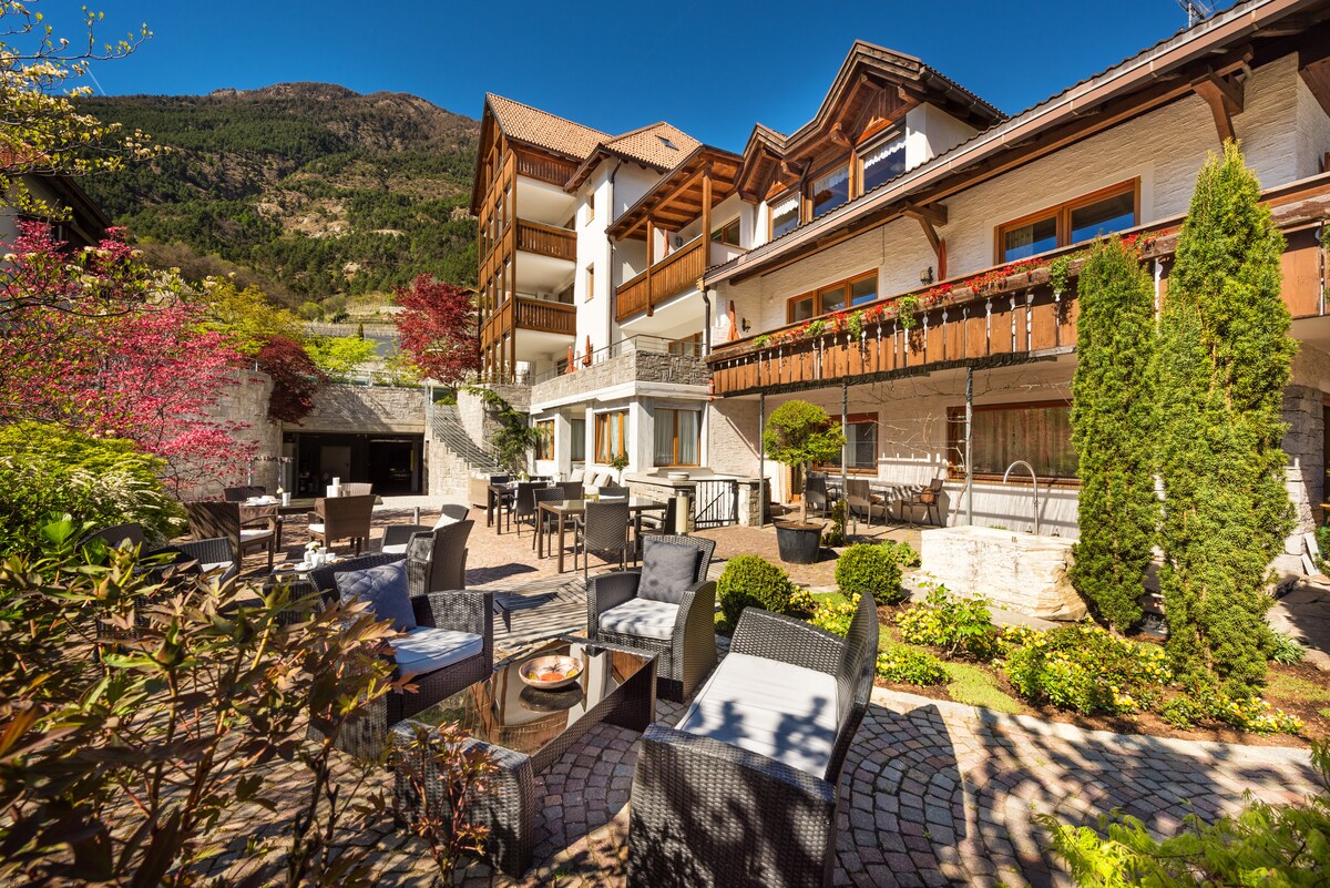 A well-maintained outdoor area is surrounded by greenery and flowering plants, featuring a variety of seating options, including woven chairs and tables. The building, with its charming architecture, offers multiple stories and balconies that overlook the serene landscape of the South Tyrolean mountains.