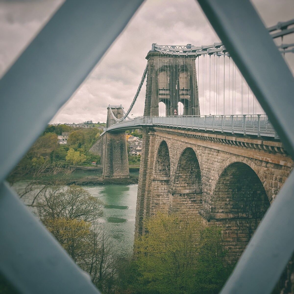 A historic stone bridge is captured spanning a river, with its tall arches and towers visible. Green trees line the water's edge, and a cloudy sky provides a muted backdrop, emphasizing the architectural features of the bridge.