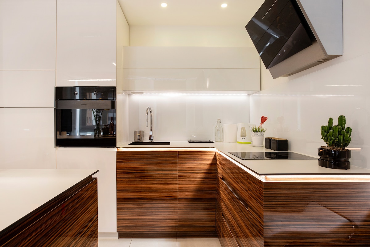 A modern kitchen is featured with sleek cabinetry in a dark wood finish. Stainless steel appliances, including an oven and a cooktop, are visible. A small potted plant sits on the countertop, and under-cabinet lighting enhances the bright, clean design.