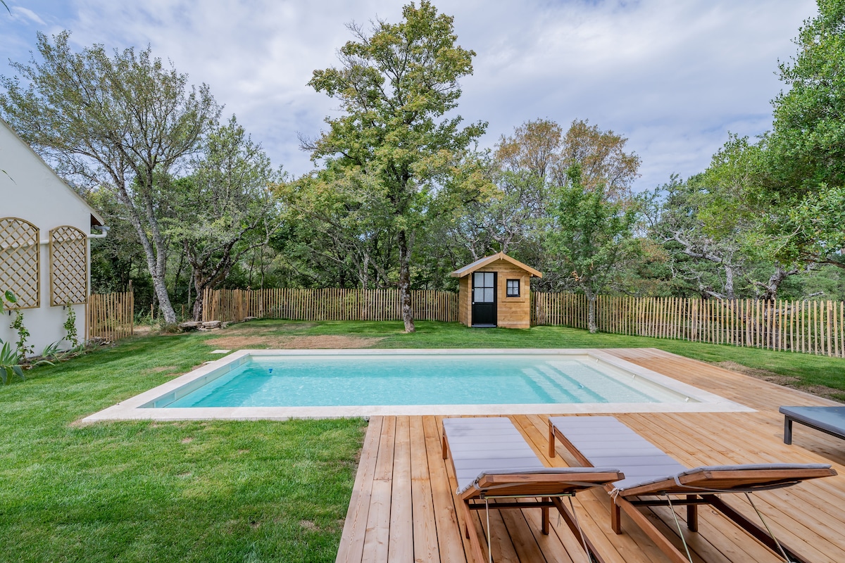 A heated swimming pool is framed by wooden decking, surrounded by lush greenery and a wooden fence. Two lounge chairs are positioned beside the pool, inviting relaxation. A small shed is visible in the background, adding charm to the tranquil outdoor space.