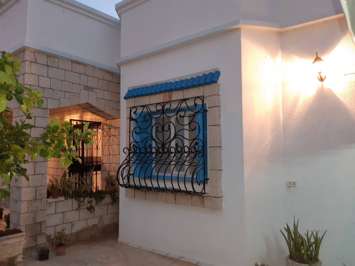 A traditional Tunisian house exterior is depicted, featuring a decorative window with ornate wrought iron bars. The window is framed in light blue, contrasting with the white plaster walls. Soft outdoor lighting highlights the entrance, creating a warm ambiance in the evening.
