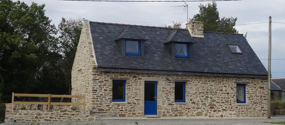 The stone exterior of a charming countryside house is highlighted, featuring a slate roof and blue accents on the windows and door. A wooden railing surrounds a small outdoor area, with greenery visible in the background.