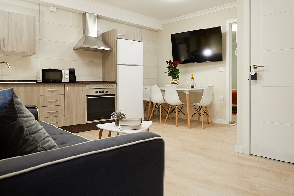 A modern kitchen area is visible, featuring light wood cabinetry, a stovetop, and a refrigerator. A dining table with four chairs is positioned nearby. The living area includes a dark sofa and a wall-mounted television, creating a functional and inviting layout.