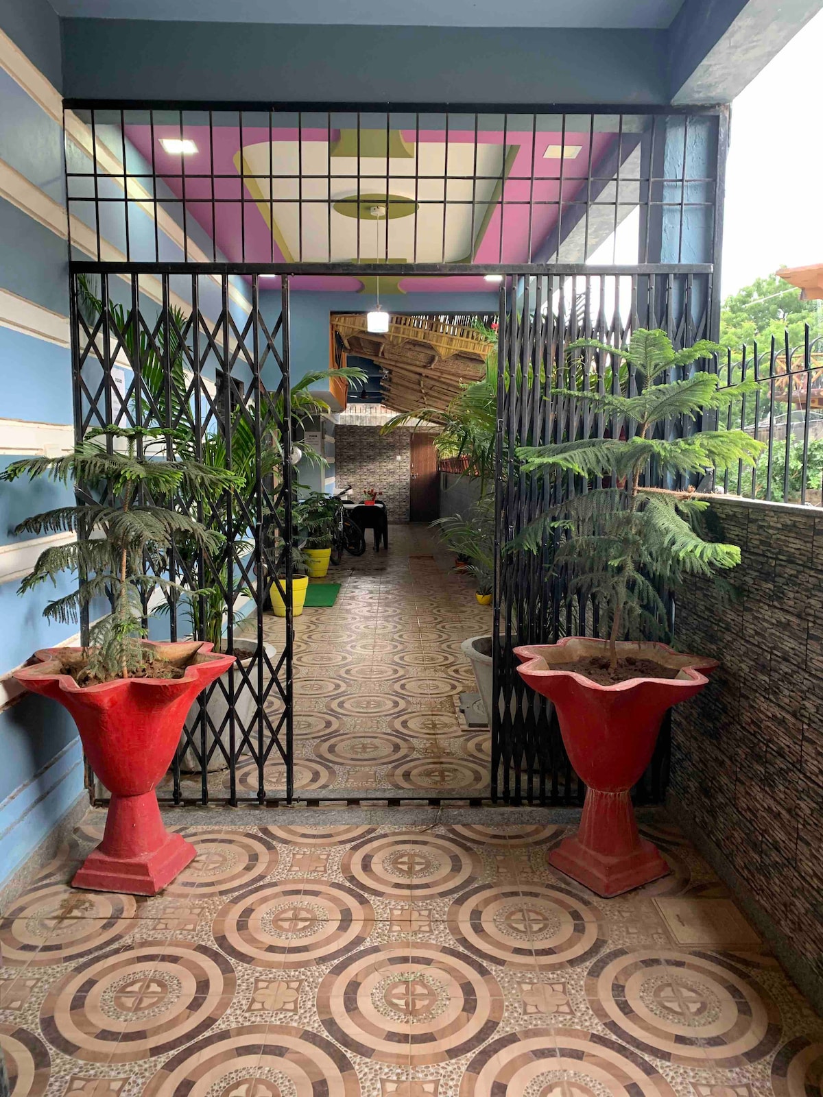 An entrance to the flat is framed by two large red planters filled with greenery. The pathway is lined with patterned tiles leading to a well-lit corridor featuring plants on either side, creating a welcoming environment.