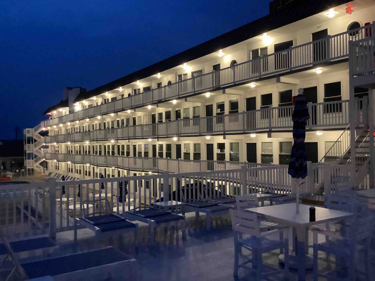 The exterior view of a multi-story building is illuminated at night by soft lighting, highlighting the balconies and walkways. A pool area with lounge chairs is visible in the foreground, offering an inviting space for relaxation.
