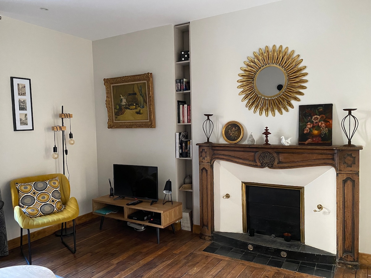 A cozy living space is featured, showcasing a decorative fireplace with an ornate wooden mantle. A round mirror is positioned above it, reflecting light. A small TV stand holds a television. An accent chair with a yellow cushion adds a pop of color, complemented by warm wooden flooring.